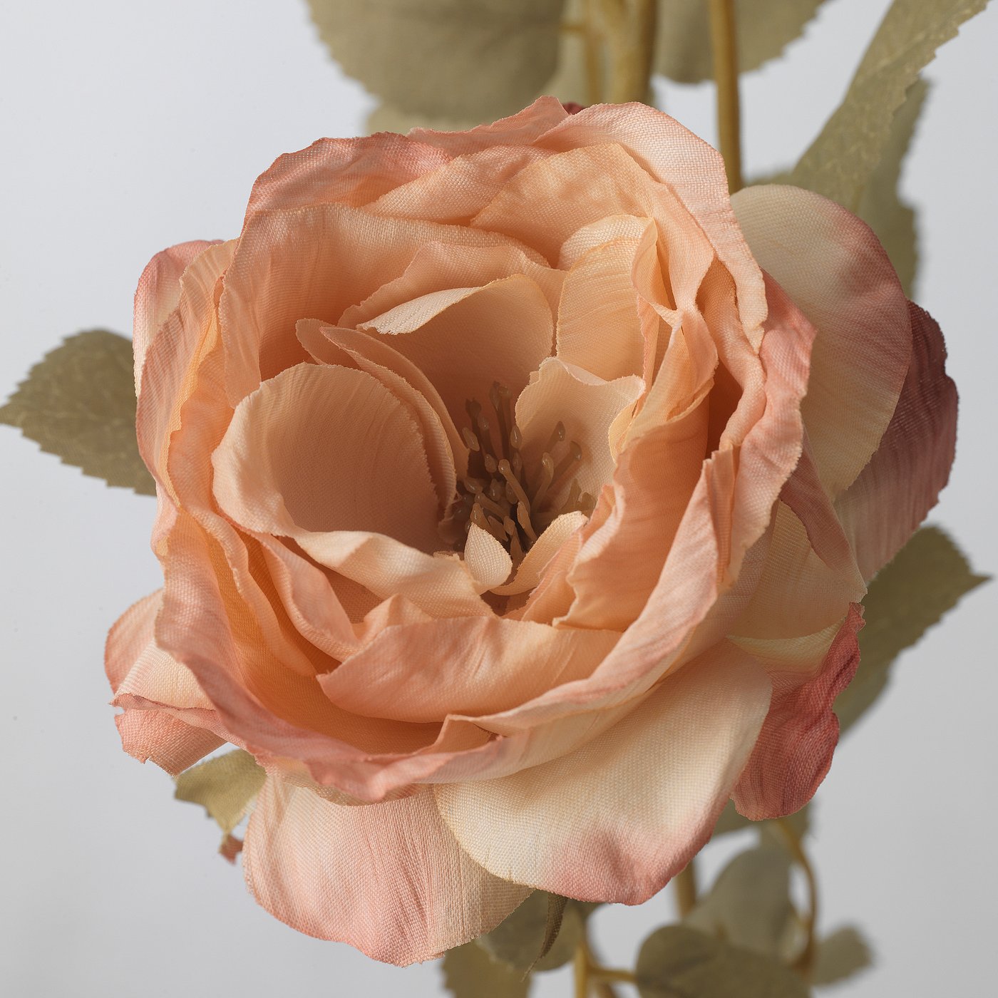 Close-up of a vibrant, realistic peach-colored artificial rose made with recycled materials. Textured petals and green leaves surround it.