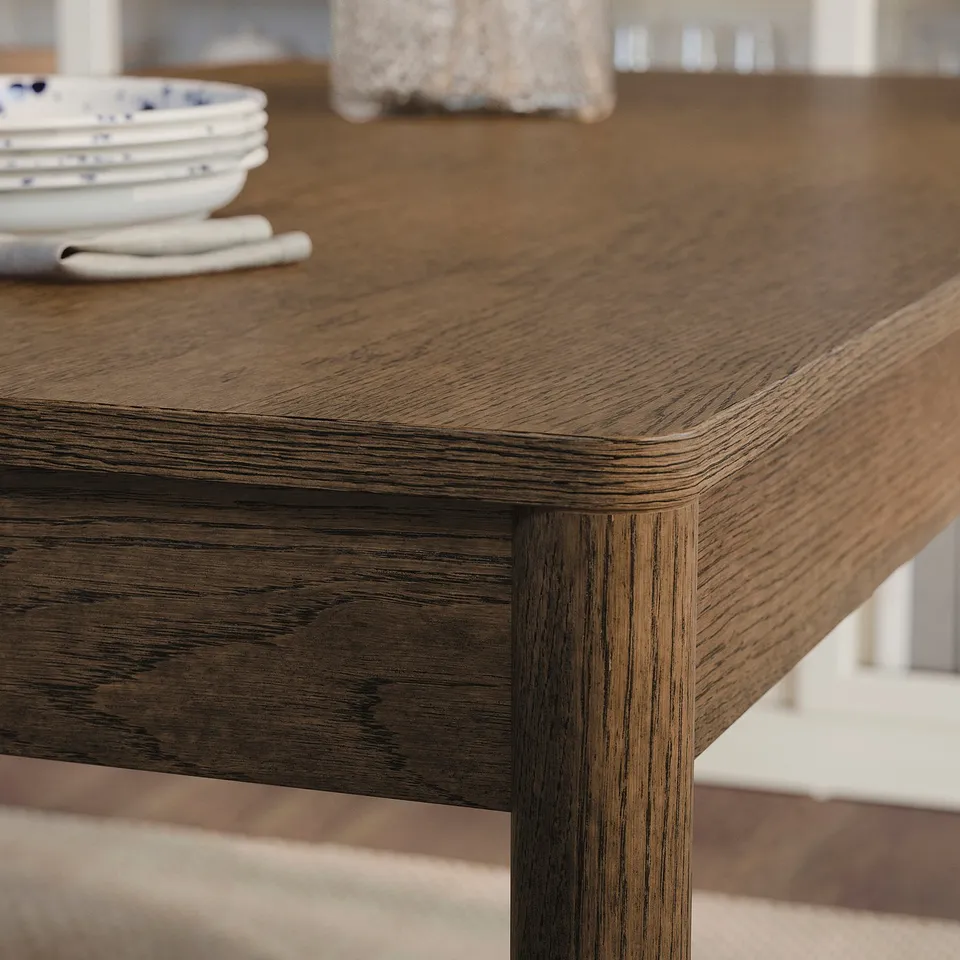 Close-up view of a wooden table with smooth, rounded edges and visible wood grain texture. A stack of plates is partially visible in the background.