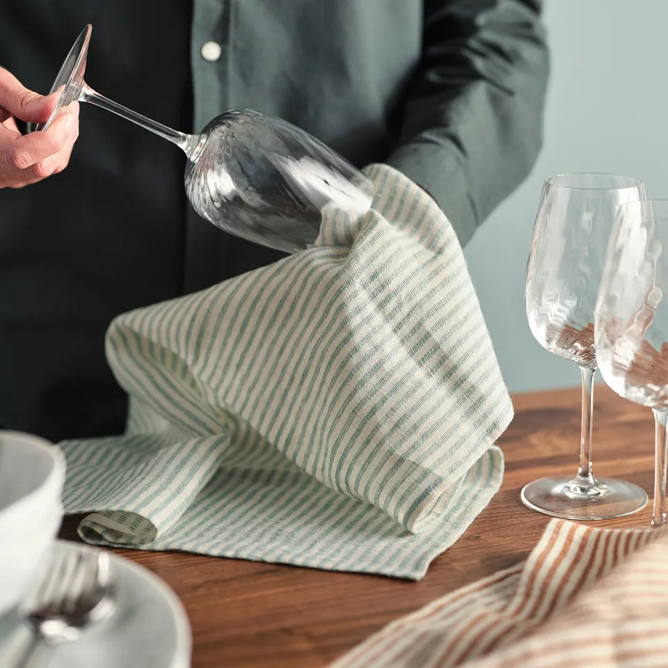 Person drying wine glass with green striped linen tea towel on wooden table. Another glass is nearby.