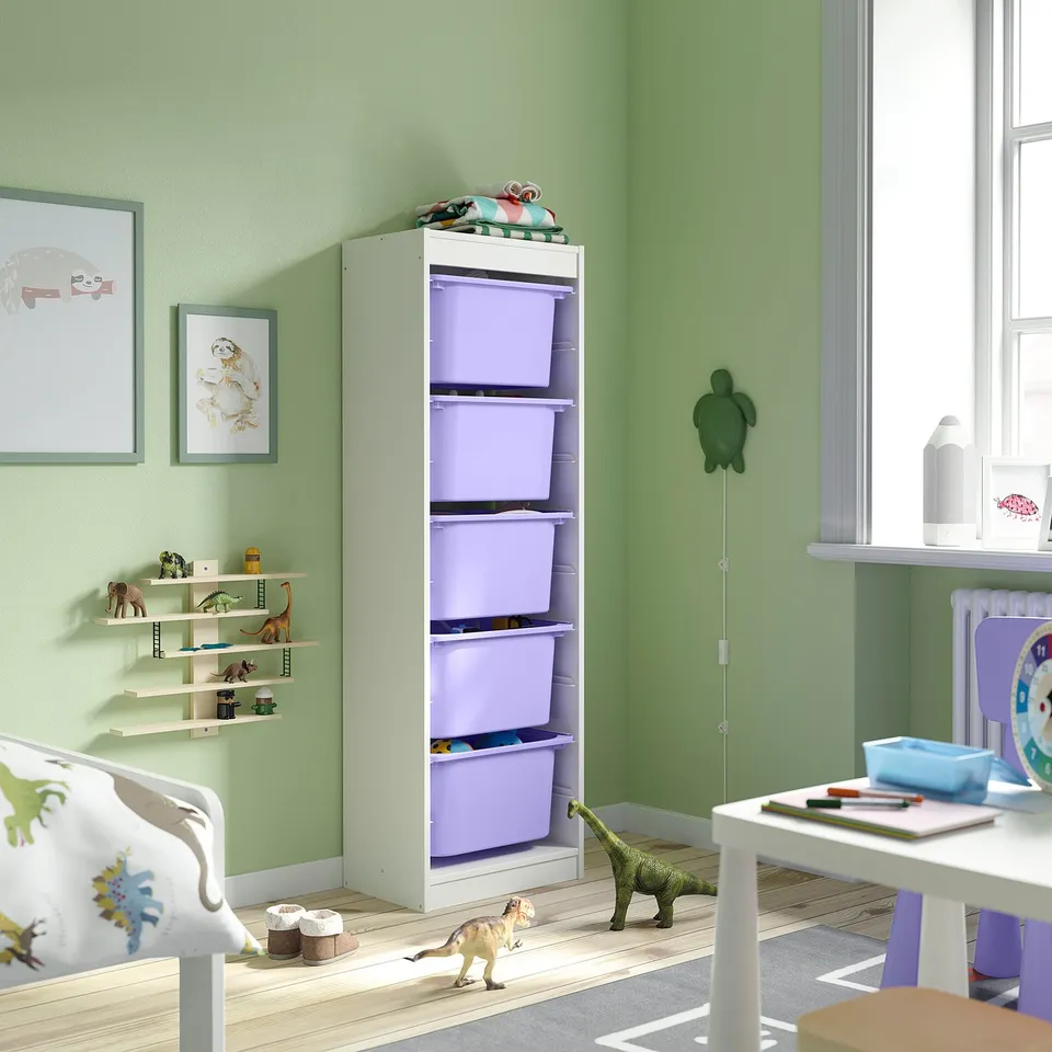 White TROFAST organiser with purple bins, standing tall beside a bed, containing childrens toys. Dinosaurs and decorative items surround it.