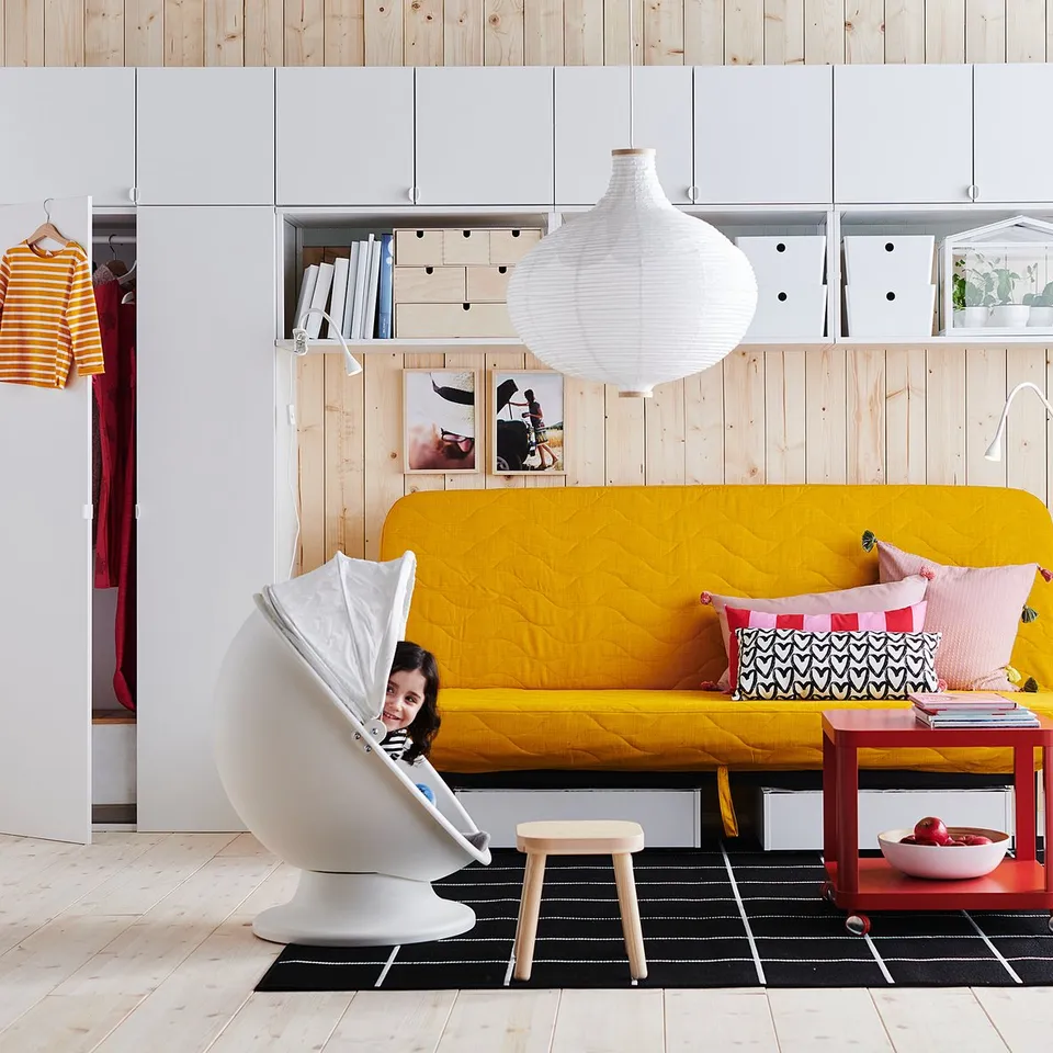 A child reads inside a white RISBYN lamp, beside a yellow sofa with colourful pillows, near a red table with fruit under a large white hanging lamp.