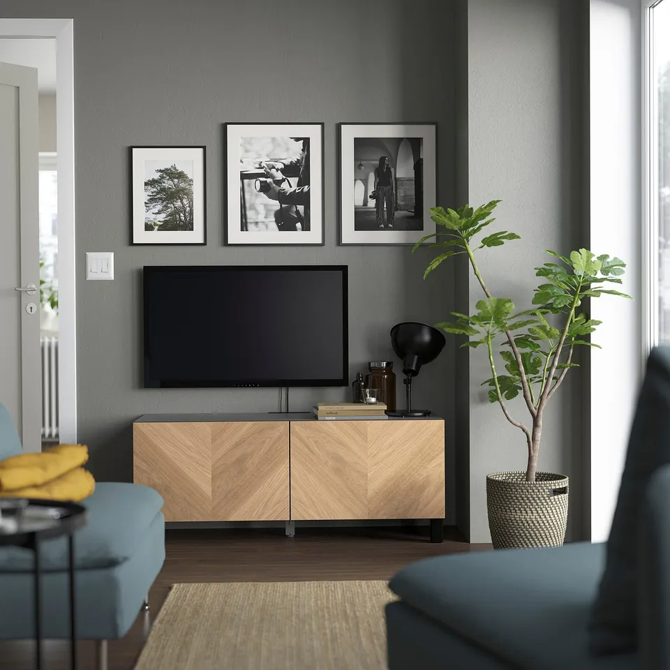 Modern living room with BESTÅ tv stand in light wood, dark grey wall, black & white pictures, flat-screen tv, and large plant. Blue & grey furniture.