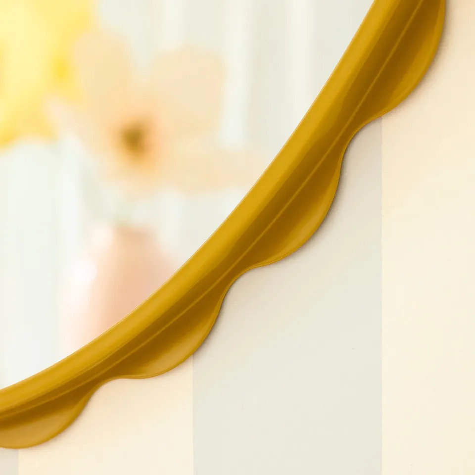 A golden mirror with a scalloped edge hangs on a light wall. The reflection is slightly blurred, emphasising the mirrors quality and design.