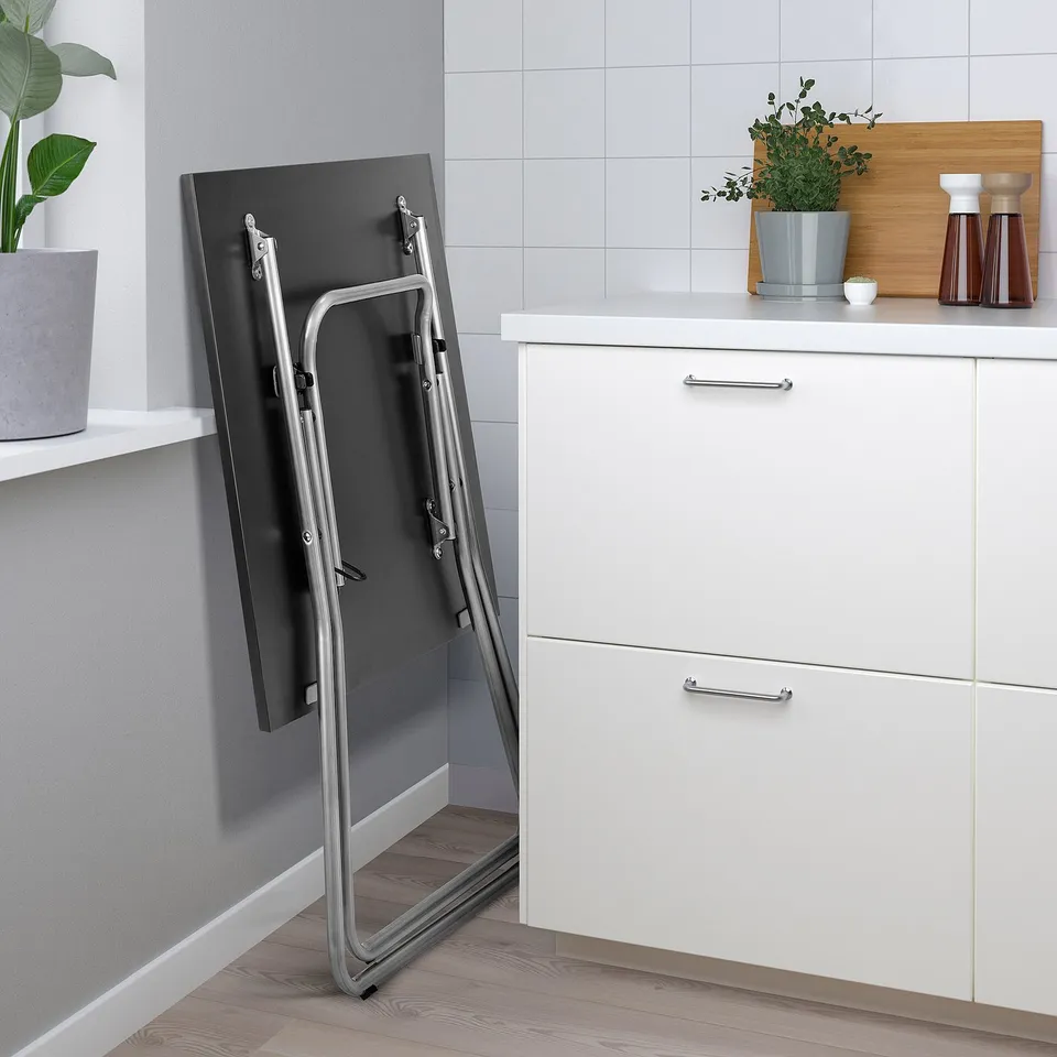 Foldable grey table leans against white cabinet, showcasing its space-saving design. White countertop holds plants and kitchen items.