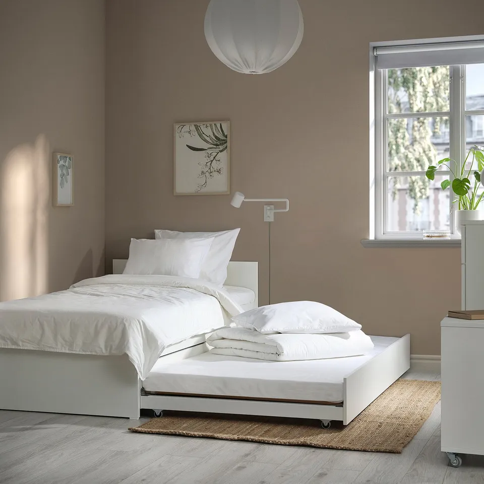 Minimalist bedroom with VIHALS white bed frame and trundle, brown wall, white linens, plant, art, lamp.