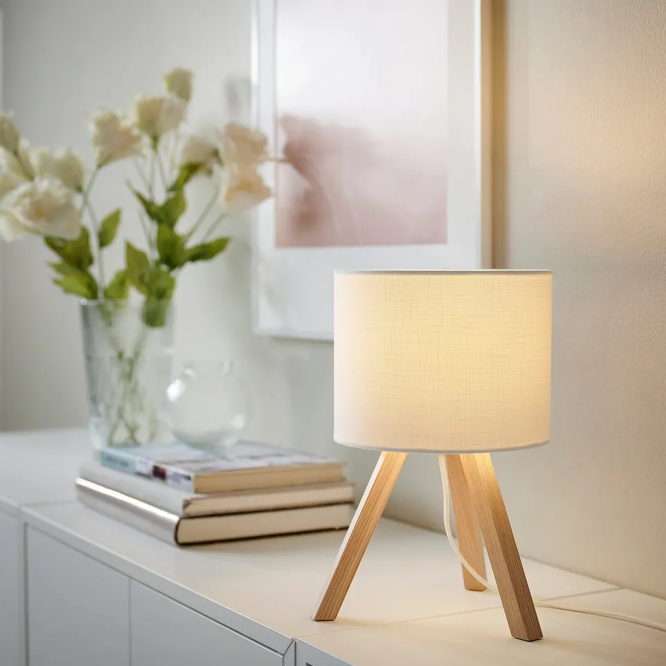 Small, modern tripod lamp STORSEGEL with light wood legs and white shade on table, beside books and vase.