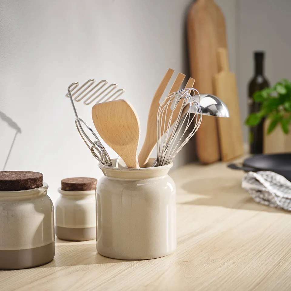Cream stoneware utensil holder with kitchen tools on wooden counter, rustic scene.