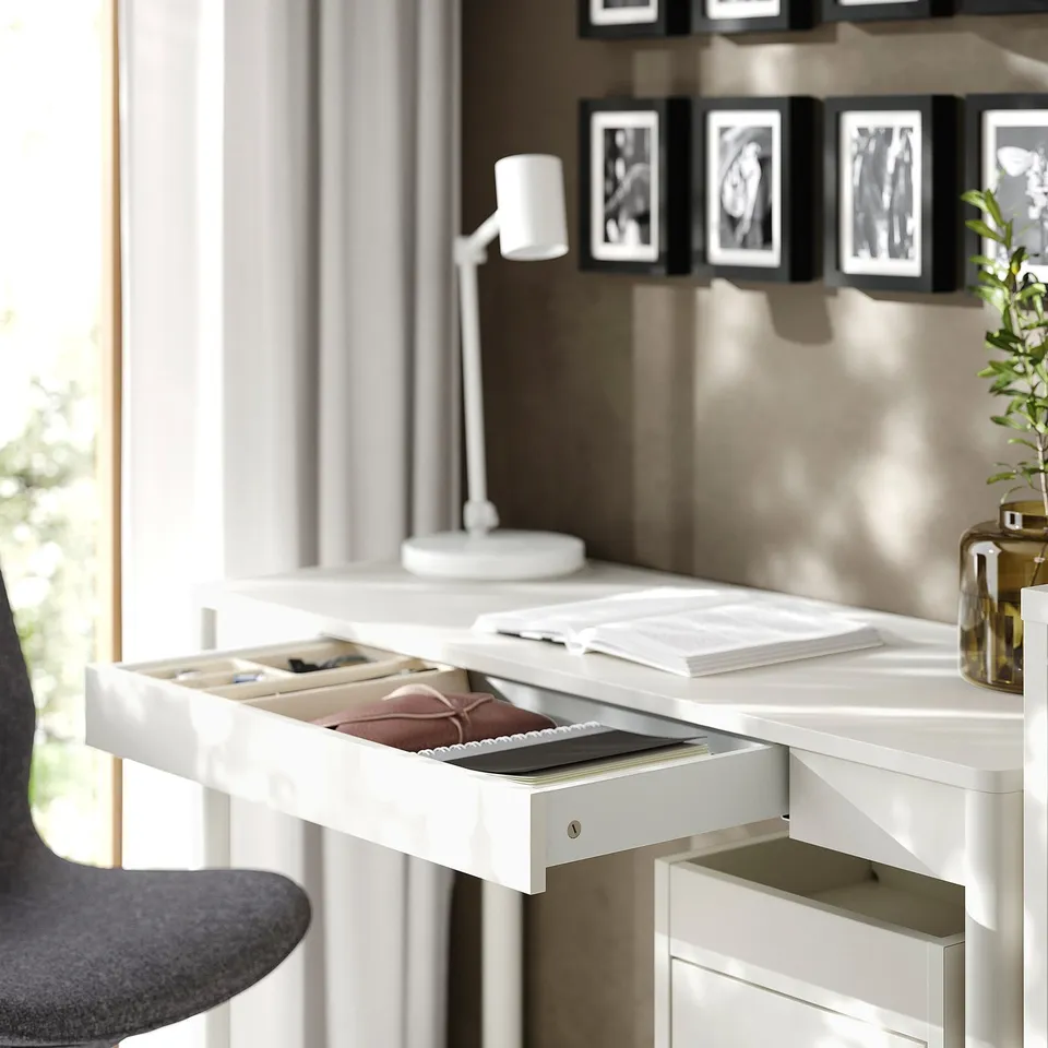 Desk with open drawer holding files, notebook, and pen near window.