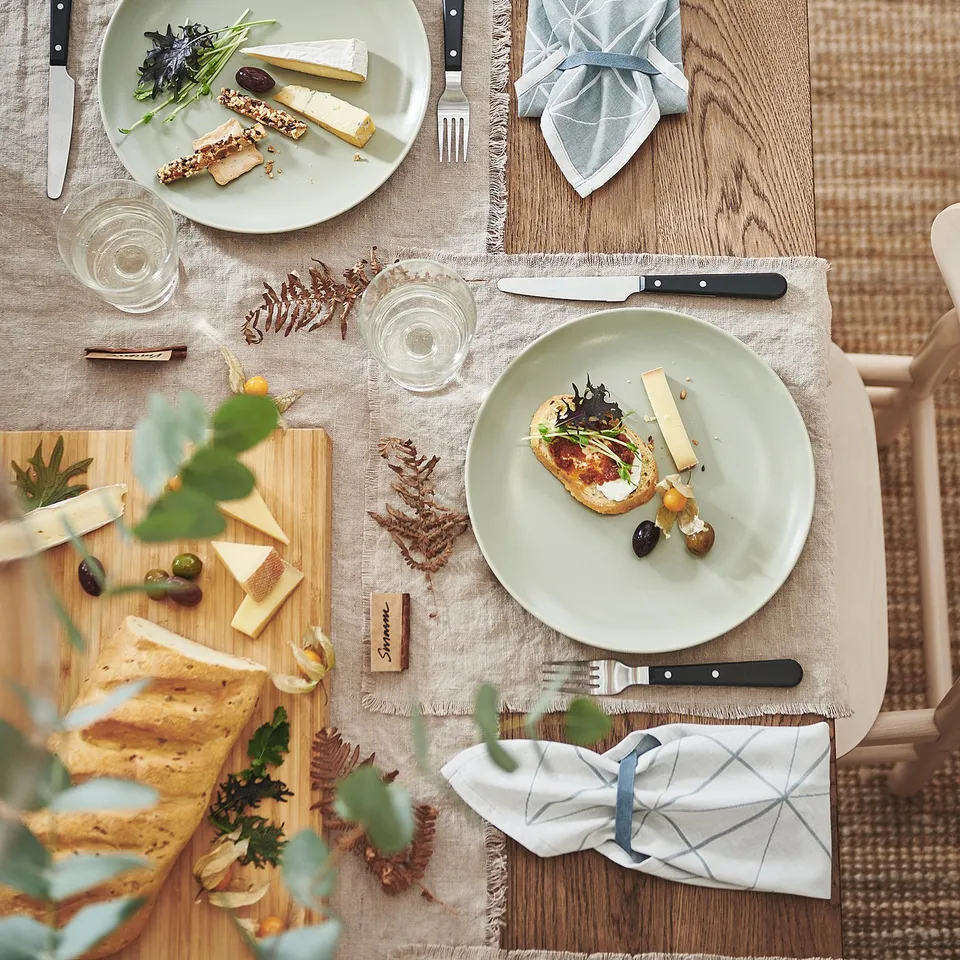 A rustic wooden table set with FÄRGKLAR white plates, grey napkins, cheese, bread, and cutlery for dining.