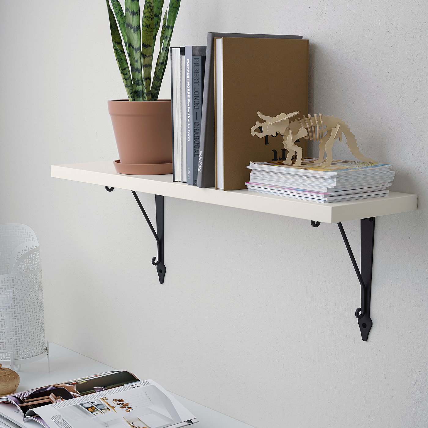 A wall-mounted shelf with black brackets holds books, a potted plant, and decorative items against a white wall, displaying various objects neatly.