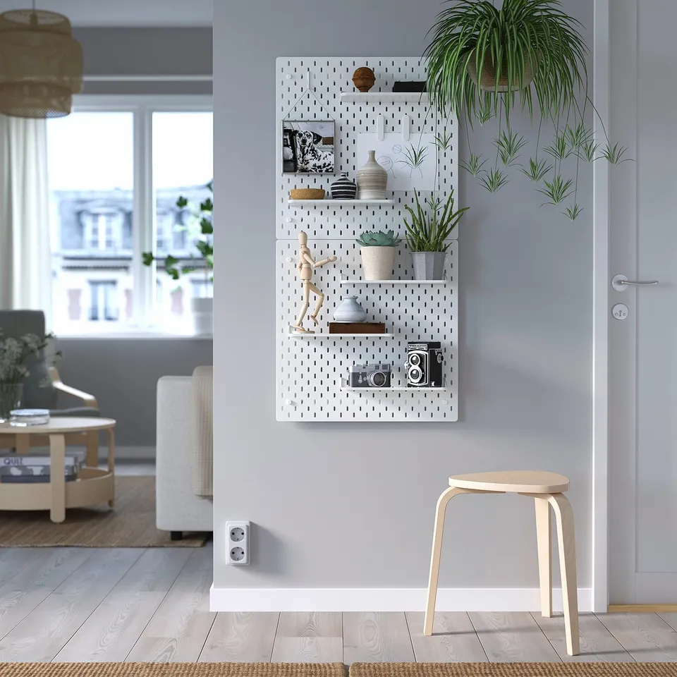 White SKÅDIS pegboard with camera, frames, plants. Wooden stool, rug, grey curtains. Living area in background.