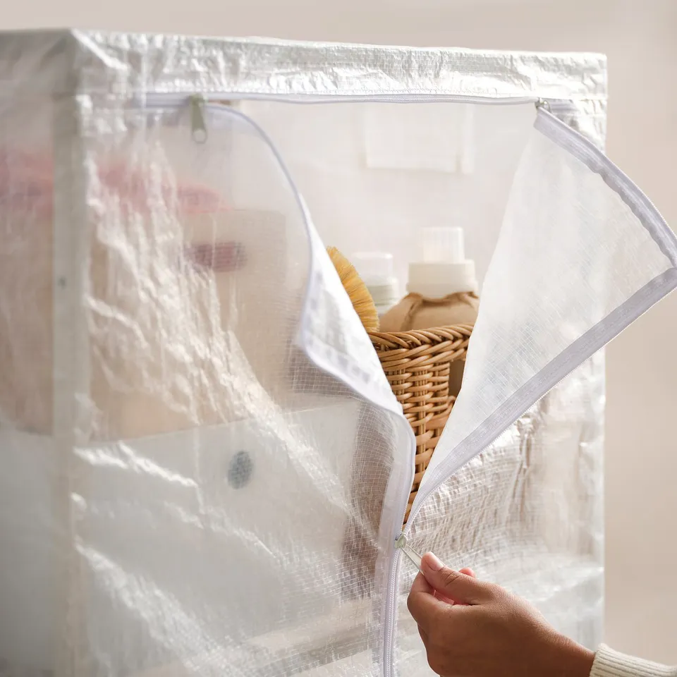 Image shows a hand zipping clear JOSTEIN storage cover, protecting items like wicker baskets and bottles inside.