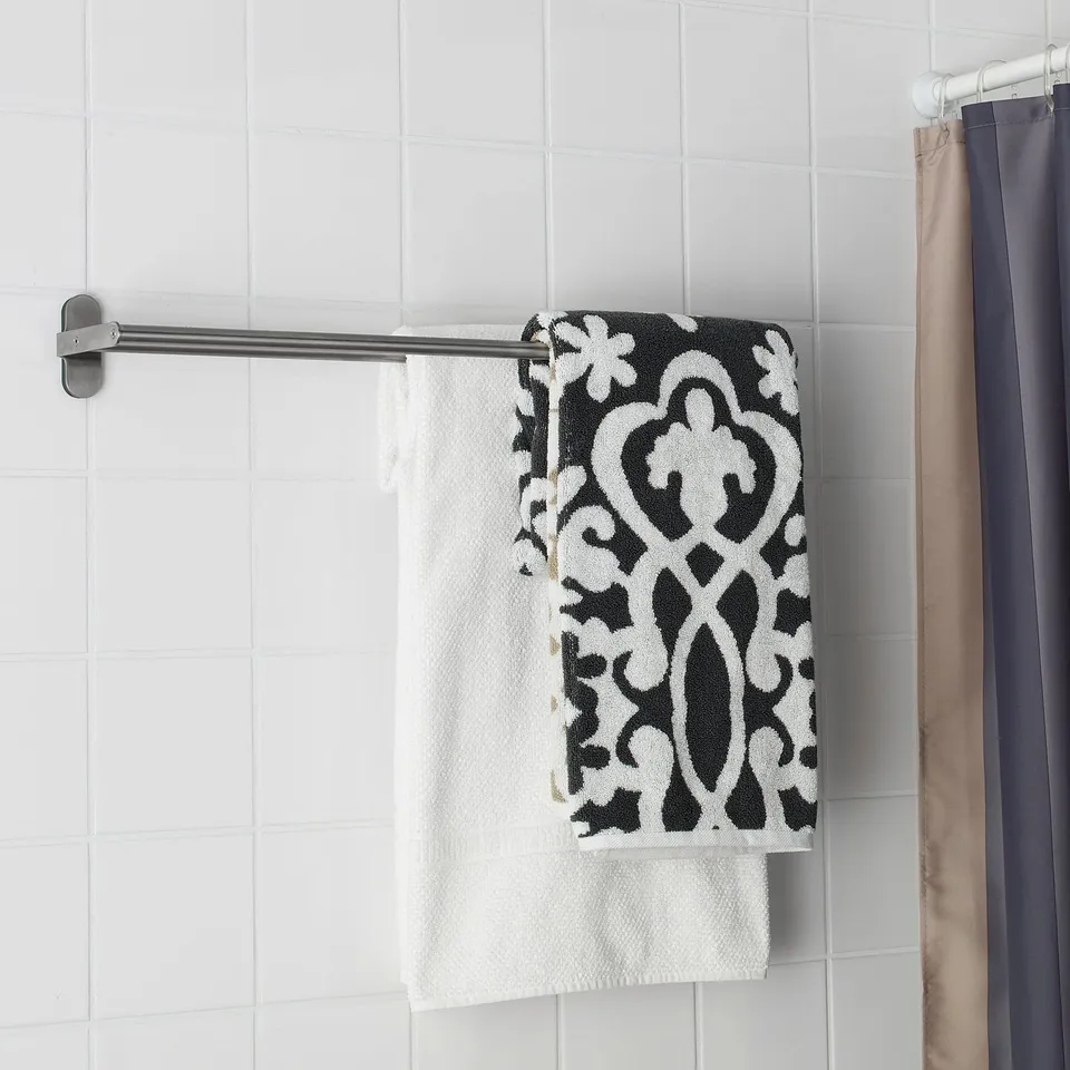 A modern silver towel rack holds two towels: a patterned black and white one on top of a plain white one, mounted on a white tiled wall.