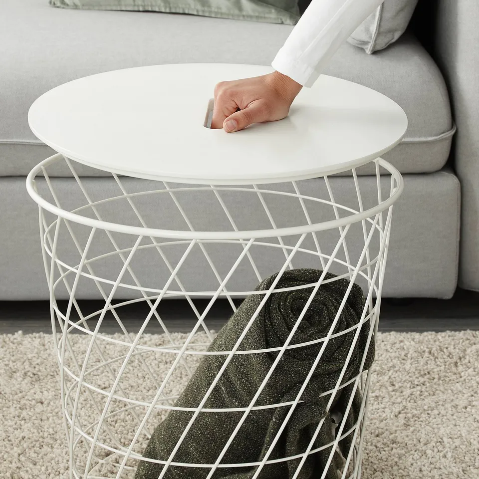 A person leans on a KVISTBRO table, which has a round white top and a wire basket base storing a green throw blanket.