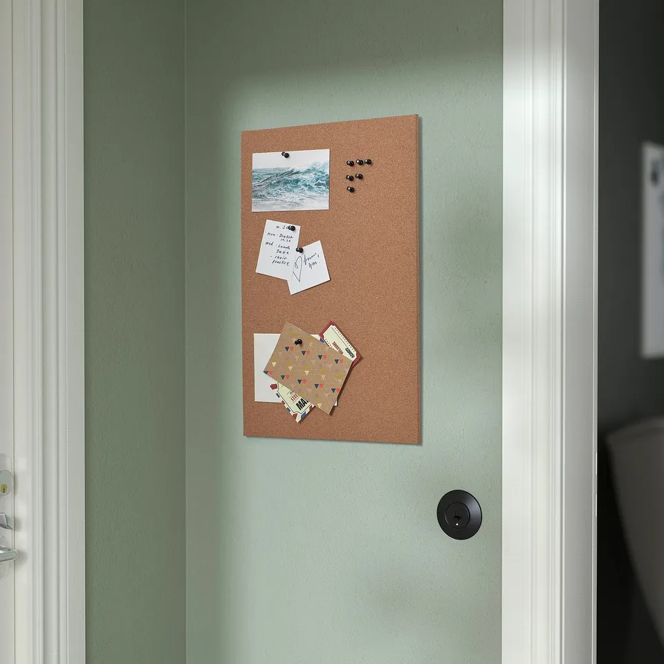 A natural cork memo board hangs on a green wall displaying photos and notes with push pins.