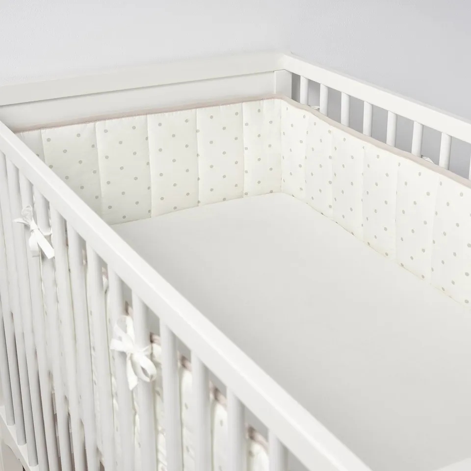 A crib with a white bumper pad that has small dots, tied with ribbons, ensuring baby safety and comfort by preventing bumps.