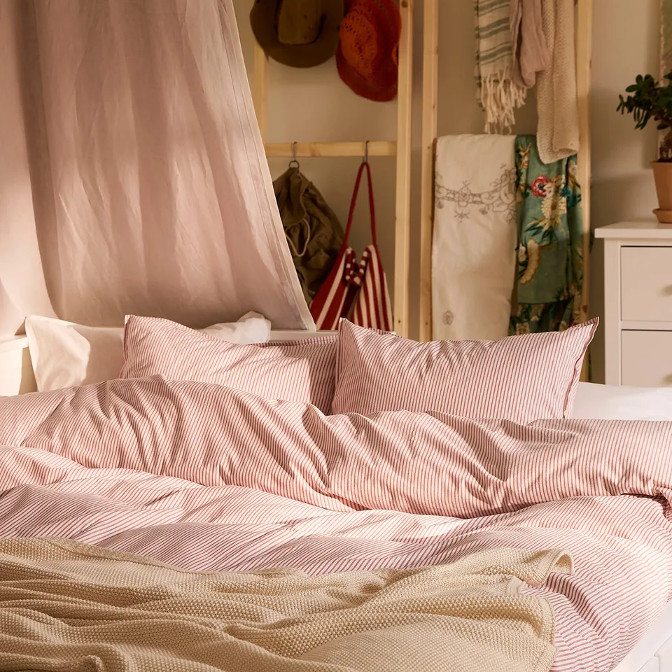 Cosy SOLFIBBLA bed with red-white striped cotton duvet, folded back, showing soft linens. Decorative shelf and hanging cloths behind.