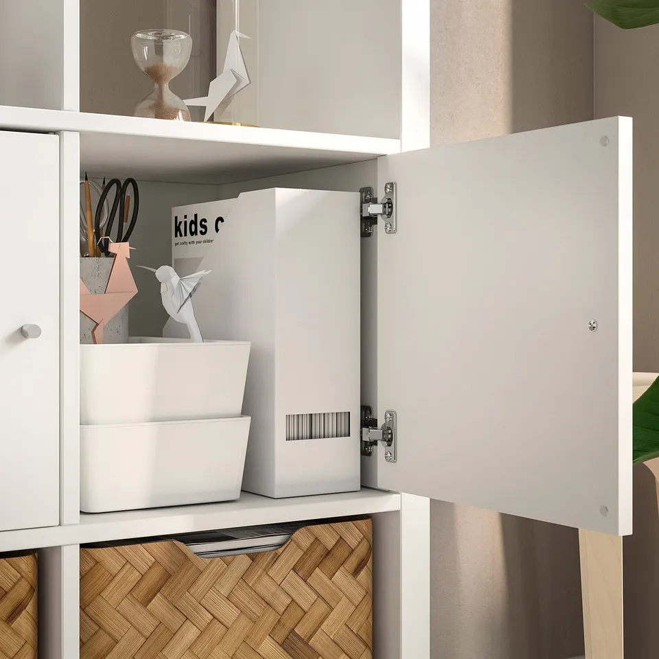 White KALLAX shelf with open door, showing organised storage including boxes, origami, and desk supplies.