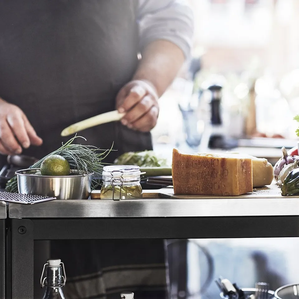 Person preparing food on a sleek black GRILLSKÄR kitchen island with stainless steel top, surrounded by cooking utensils and ingredients.