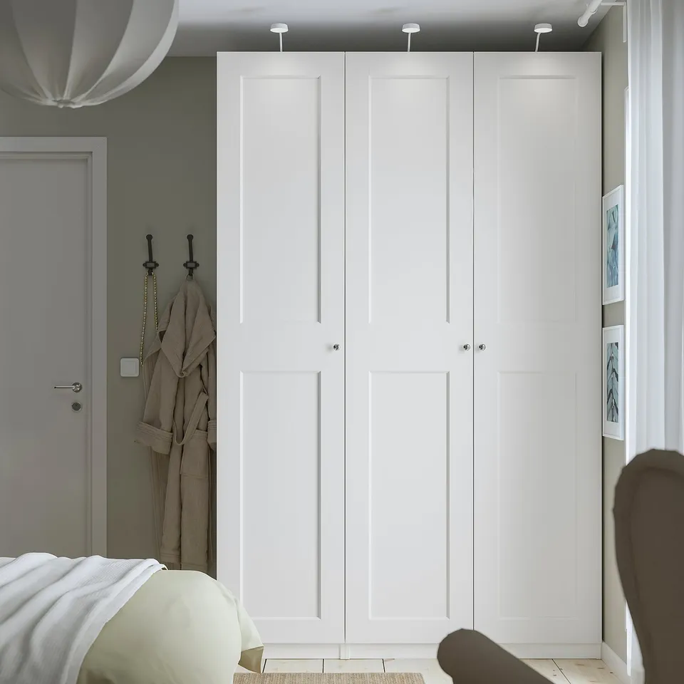 Tall, narrow white PAX wardrobe against sage green wall, with sleek knobs & grey door nearby. Dwarfs other items, showing large capacity.