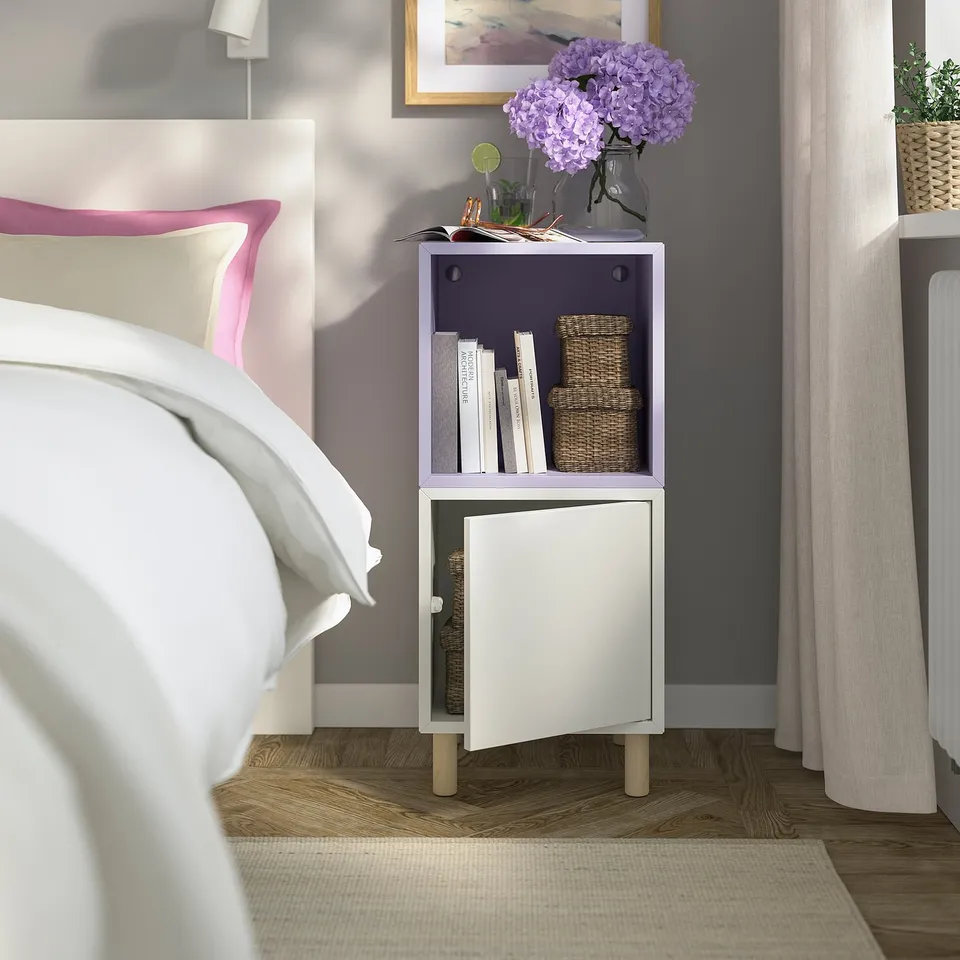 Small white EKET bedside table with a purple upper section, placed beside a bed, holding books, a vase of purple flowers, and decorative baskets.