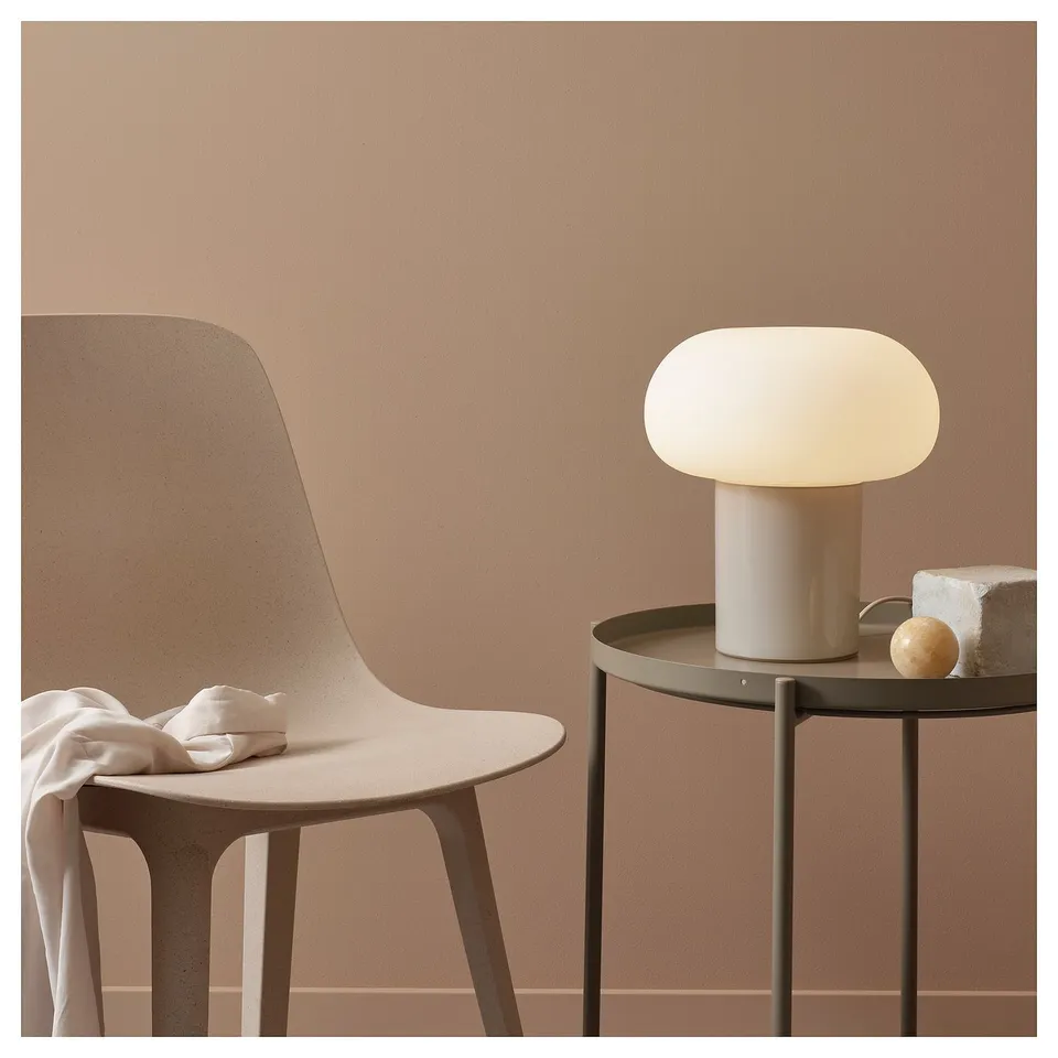 Minimalist desk setup: DEJSA lamp with opal glass shade, beige chair, round metal table, neutral tones, warm lighting.
