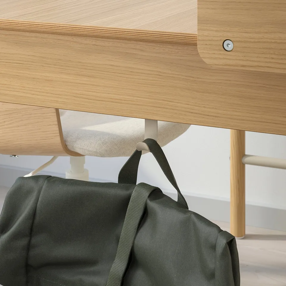 A light wood desk with a hooked dark green bag. The bag hangs from a table edge hook, demonstrating its practical use for organising accessories.