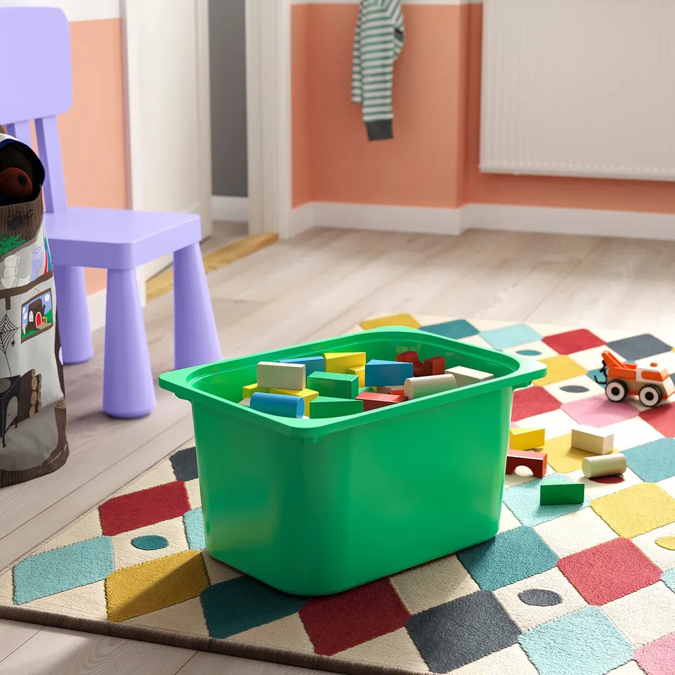 Green TROFAST storage bin full of multicoloured blocks on a geometric rug in a childs playroom.