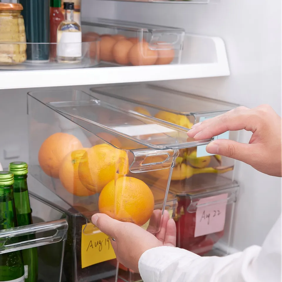 Hand places oranges into clear, stackable fridge box with lid and handle, organising fridge contents.