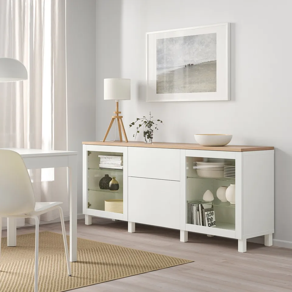 Modern white BESTÅ sideboard with glass doors. Holds dishes, books. Tall, elegant, clean look. Wood top, white paint,, under a large wall frame.