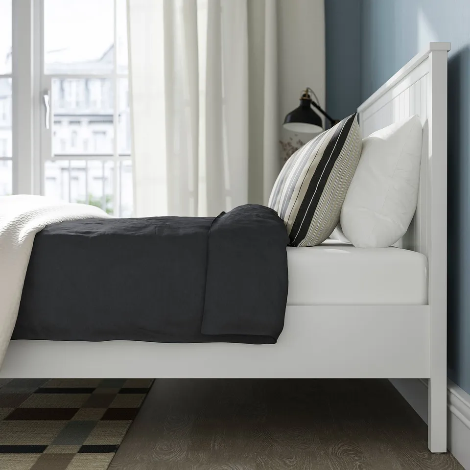 Clean modern bed with white headboard, dark grey blanket, striped pillows, white curtains, and wooden floor.