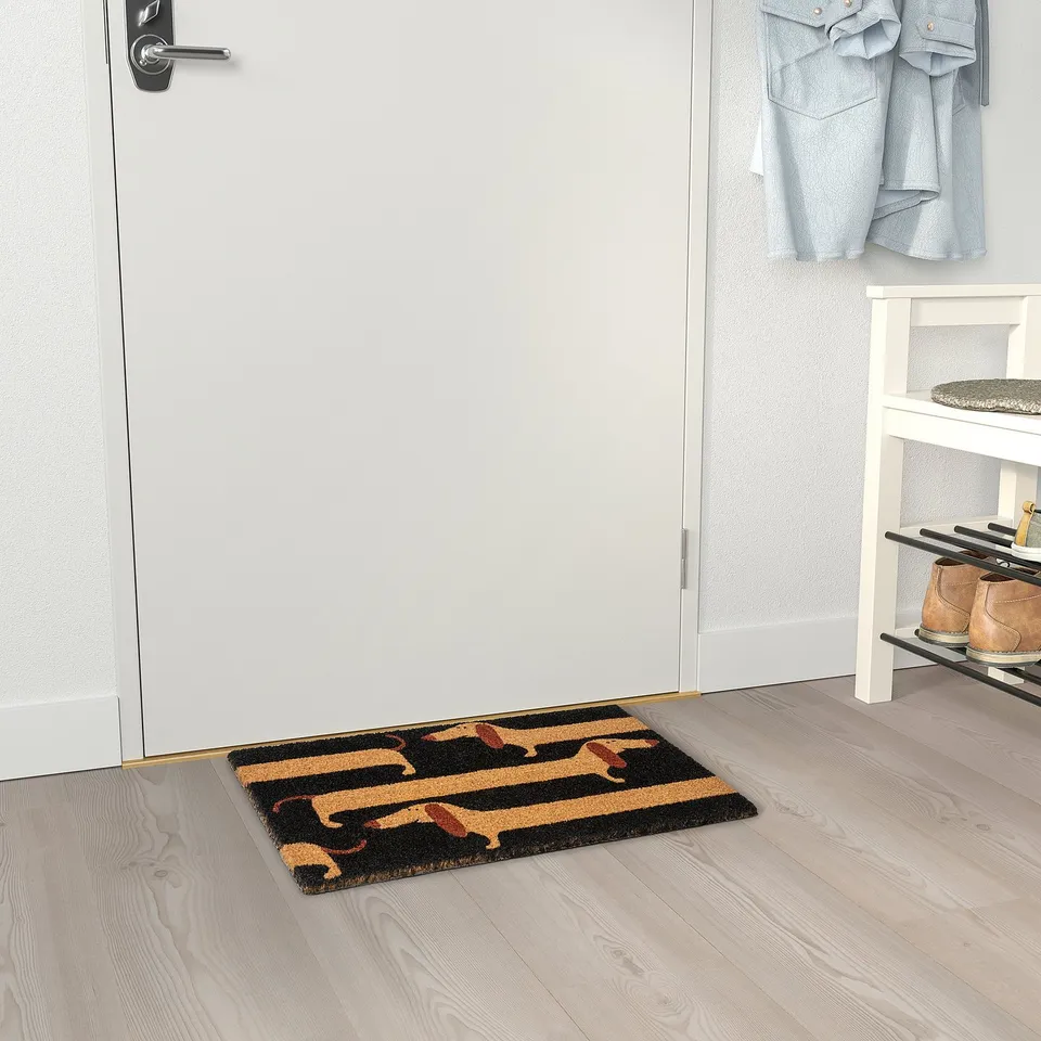 Doormat with dog patterns by entrance, preventing dirt from entering home. Black and beige mat with dachshunds lies by white door.
