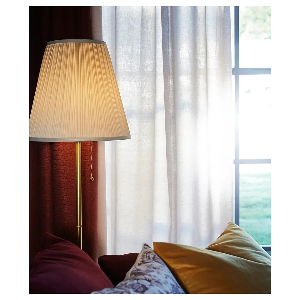 A stylish lamp named ÅRSTID with a beige shade and gold base, creating a warm atmosphere near a window.