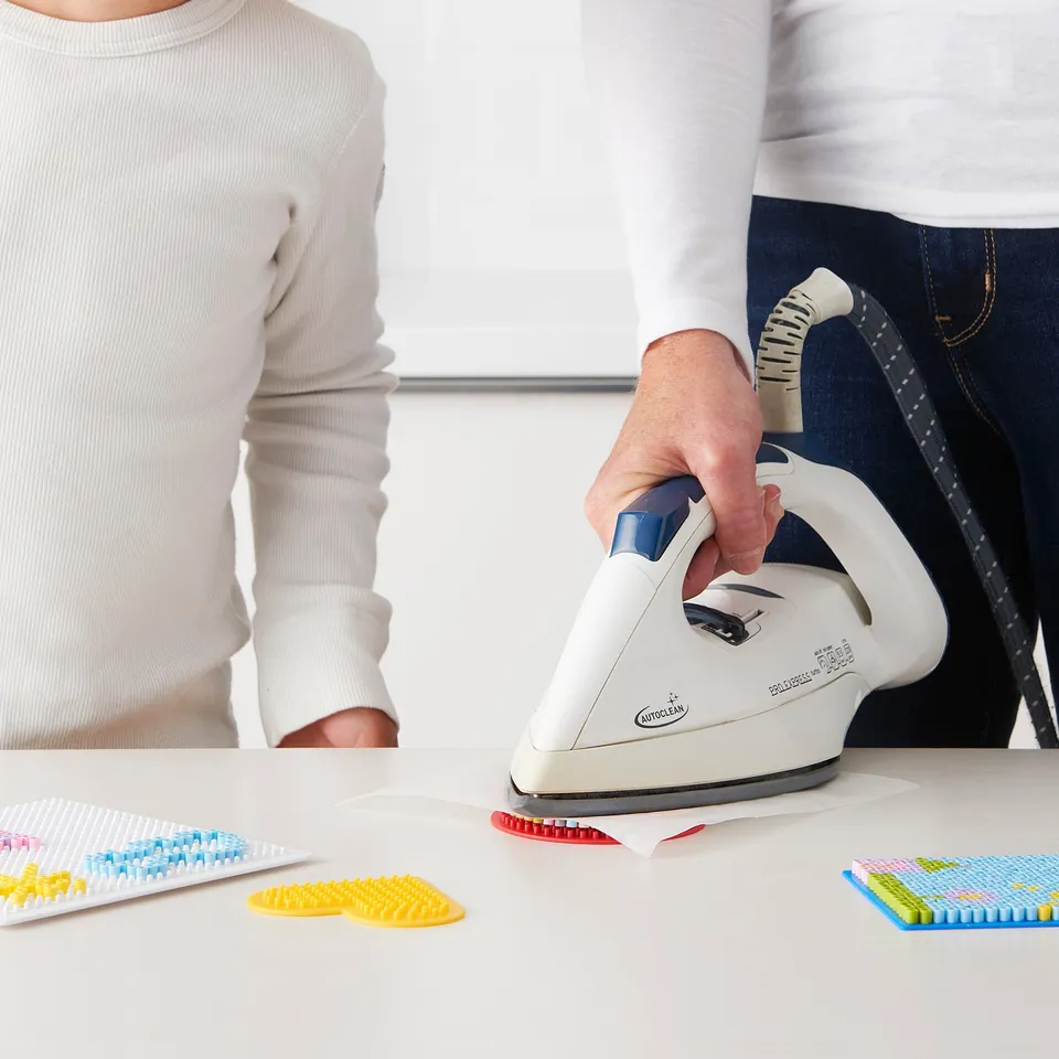 Person holding a kyssla ironing tool, creating bead art with colourful designs on a table.