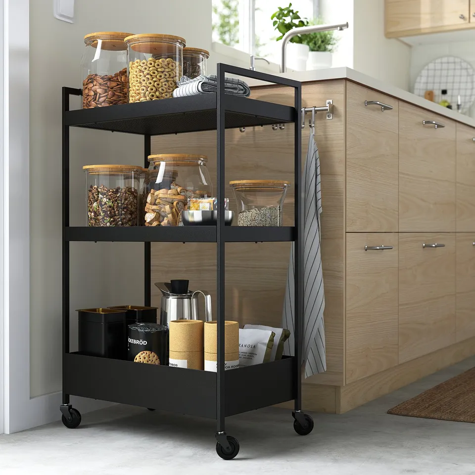 Black NISSAFORS kitchen trolley with three shelves, holding food in jars and utensils, fitting in small spaces with four wheels.