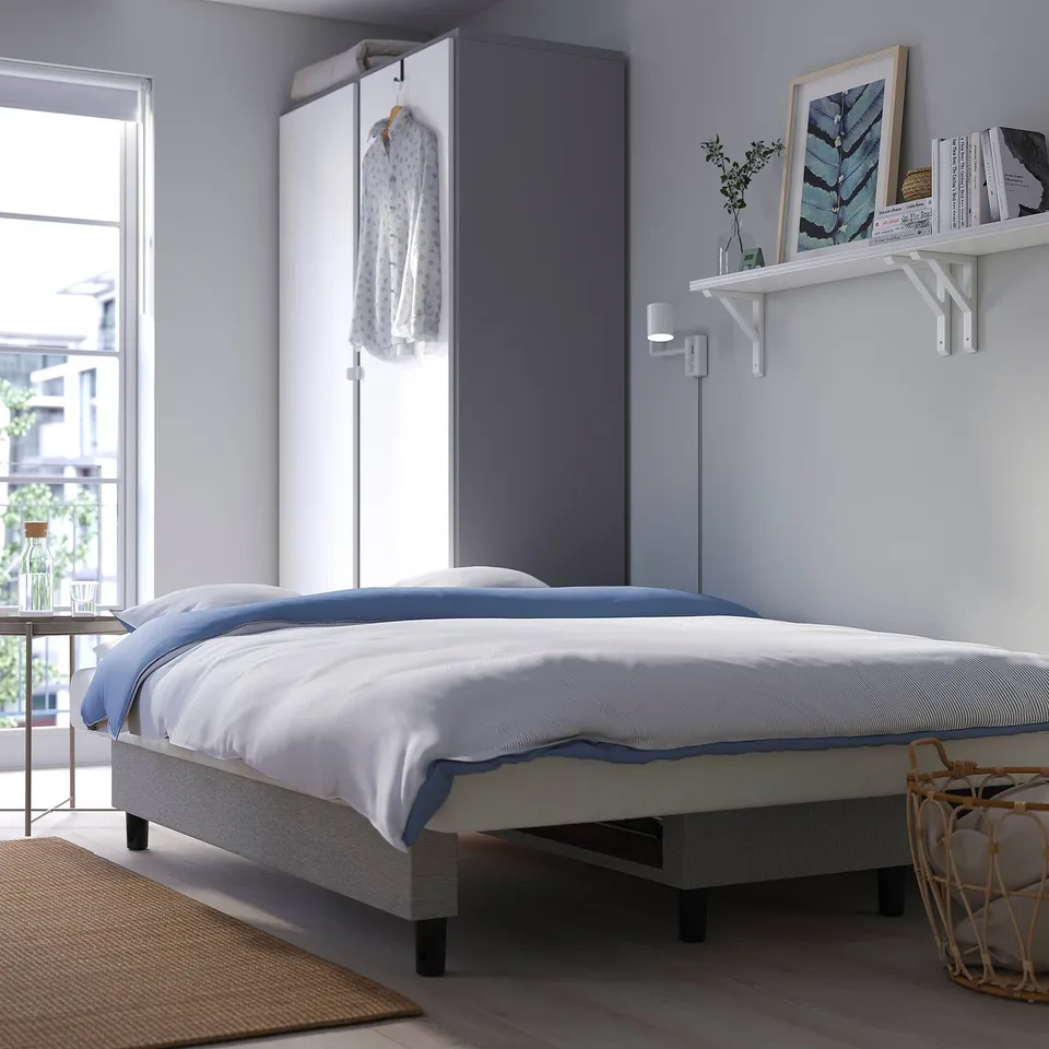 Modern bedroom with a white bed, blue comforter, wall-mounted light, and shelf with books.