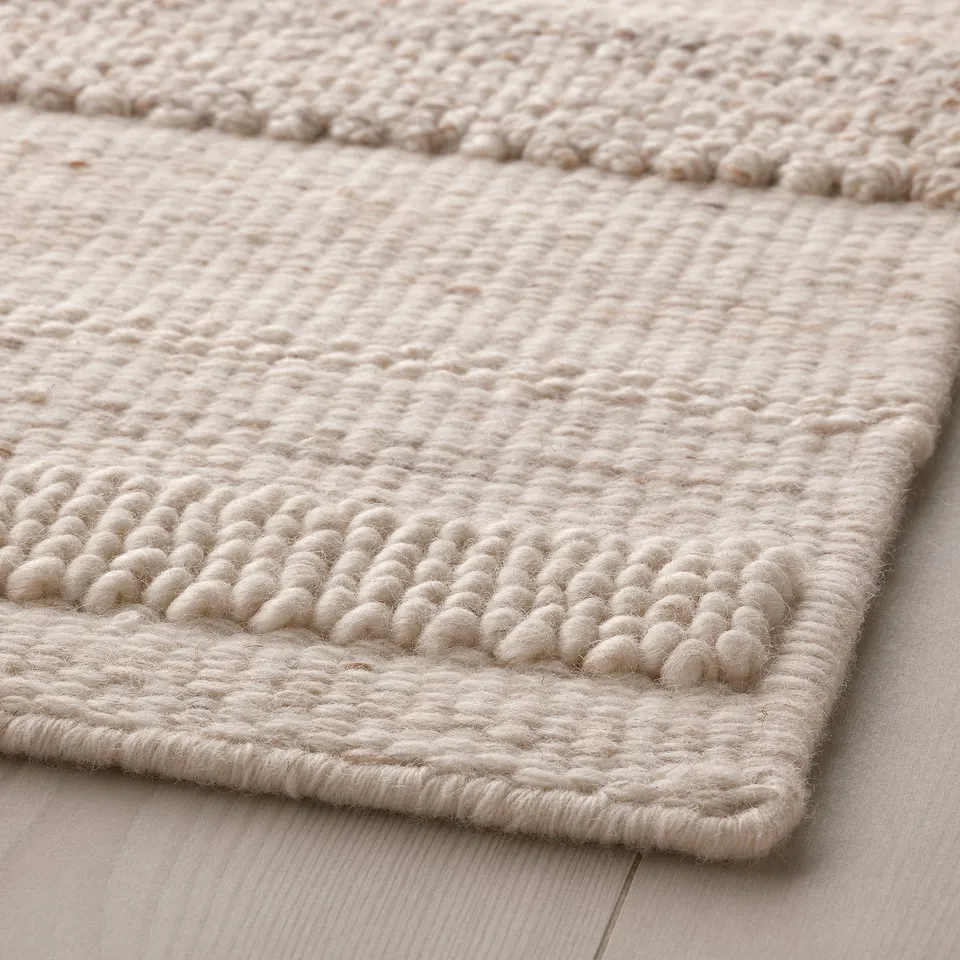 Soft, intricately woven beige rug on a wooden floor, showcasing a blend of textures and patterns, exuding warmth and craftsmanship.