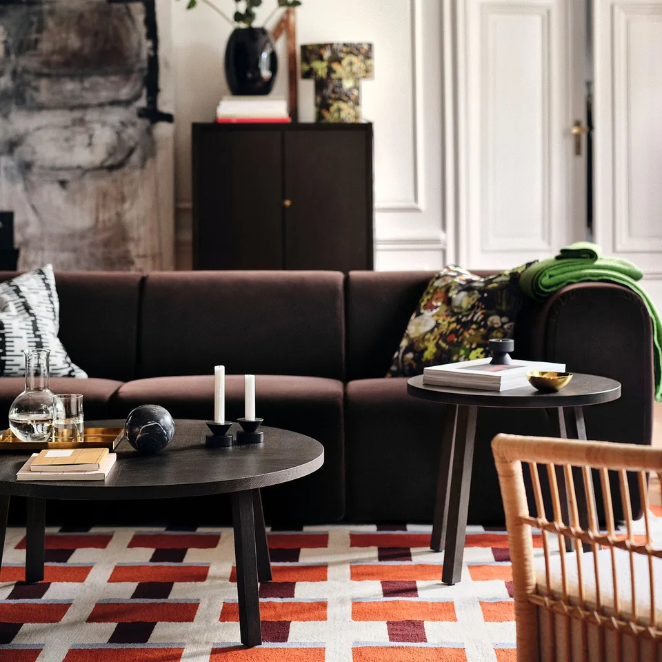 A stylish living room with a dark brown sectional, colourful geometric rug, STOCKHOLM 2025 in reds and oranges.