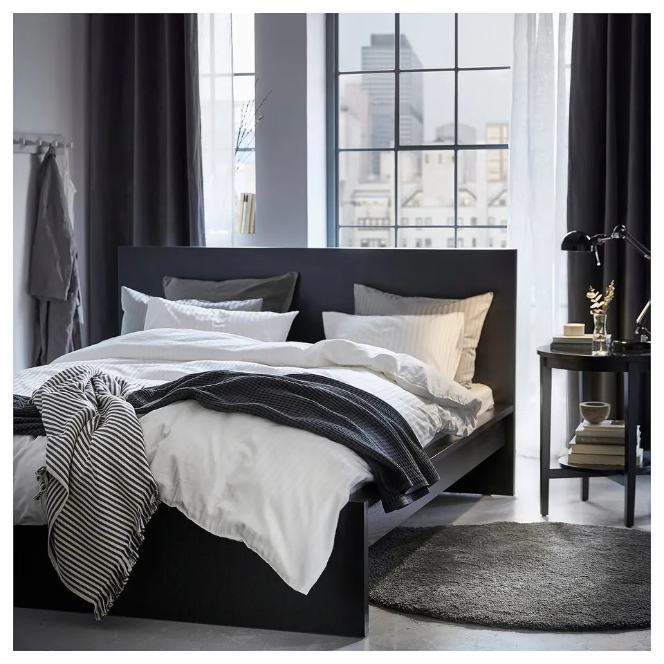 Bedroom with large windows, black VIDGA bed with white bedding, striped blanket, bedside table, lamp, and city view.