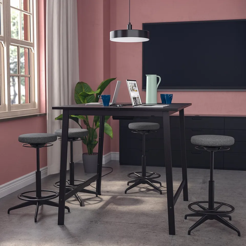 MITTZON high-conference table, black, steel/wood, rectangular, with four adjustable stools, a potted plant, laptop, cups, and a large screen behind.