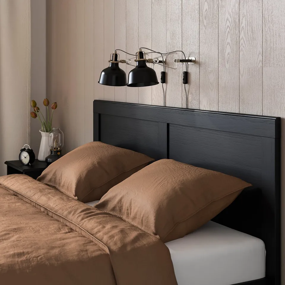 Brown bedding on dark wood bed, two black lamps, bedside table with vase and clock.