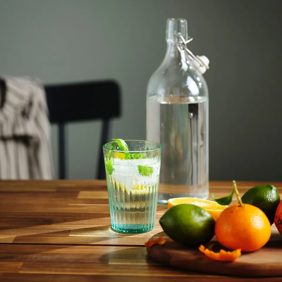 A clear glass filled with water and a mint leaf sits on a wooden table, flanked by limes and oranges. A glass bottle of water is also present.