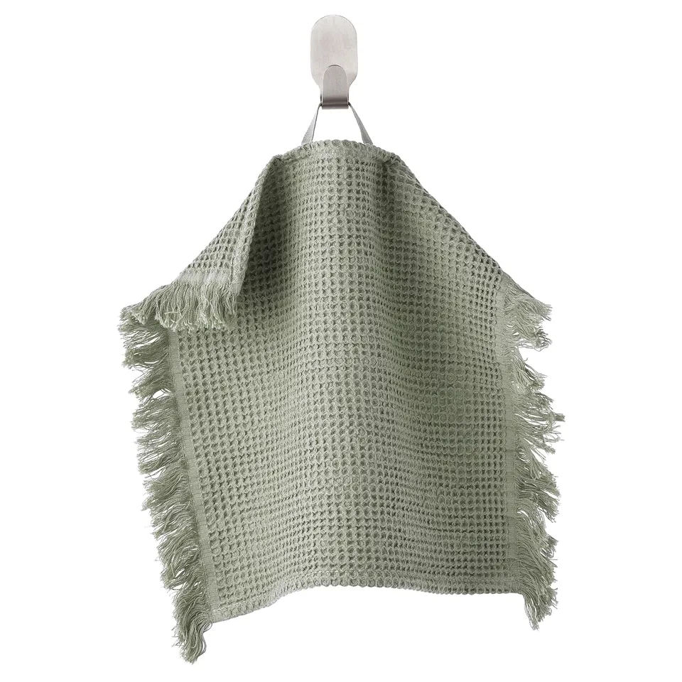 A green VALLASÅN towel hangs on a hook. Made of cotton and viscose, it has fringe on edges and a textured weave.