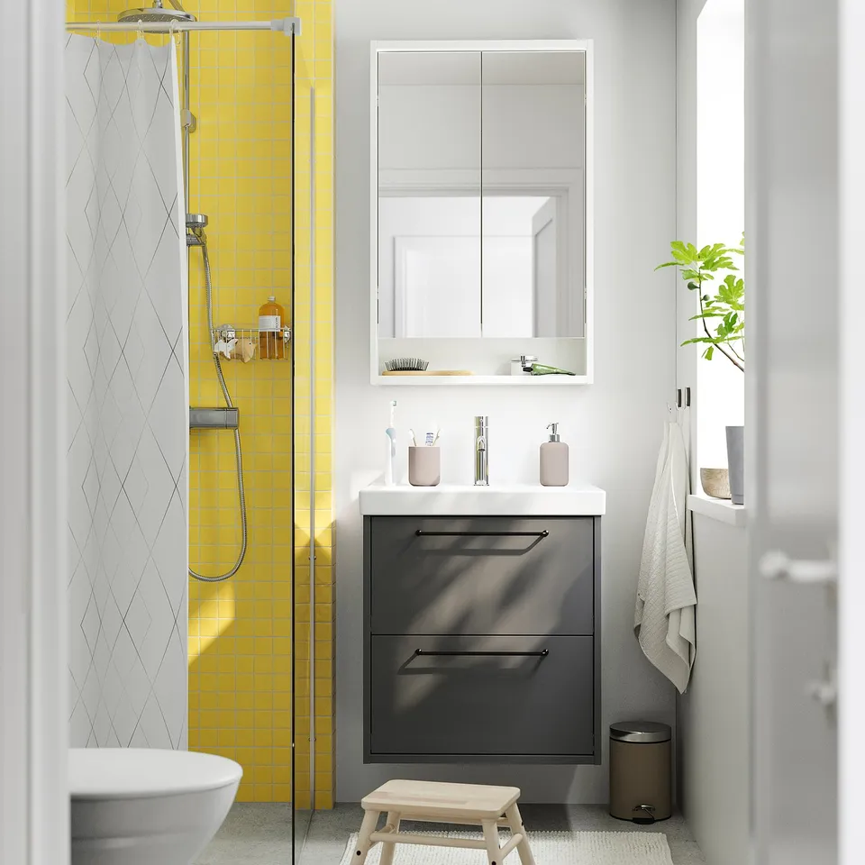 Contemporary bathroom with dark grey HAVBÄCK vanity, yellow tiled shower, white sink, mirror cabinet, and wooden stool.