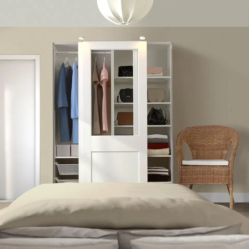 Organised bedroom with white PAX wardrobe. Glass doors showcase clothes, blending with neutral bed tones and wicker chair.