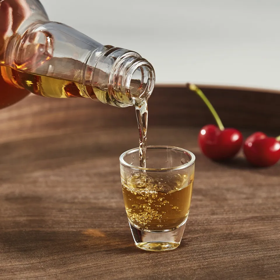 A transparent glass is filled with liquid from a bottle, likely used for tasting strong drinks like baijiu or sake. The glass is on a wooden table.