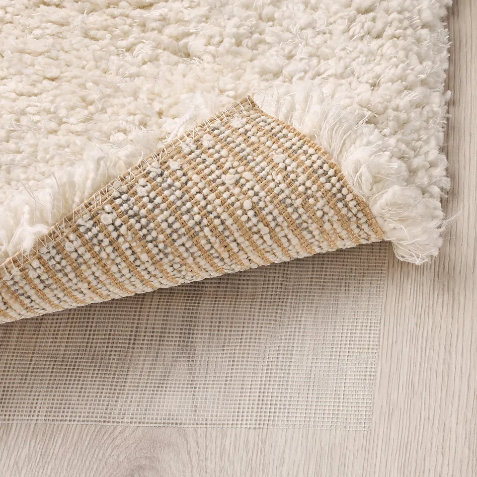 Fluffy white rug, folded corner, revealing thick, textured weave and anti-slip mesh underside for safety and comfort.