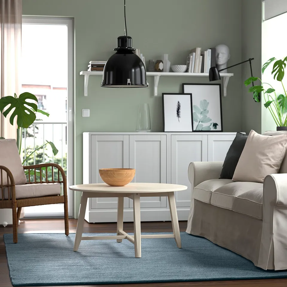 Modern living room with gray-blue TIDTABELL rug, wooden coffee table, beige couch, wicker chair, black pendant lamp, and green wall.