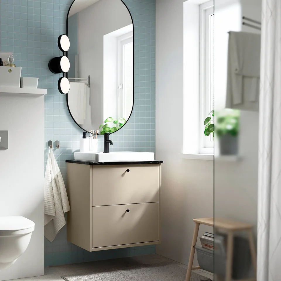Modern bathroom with beige HAVBÄCK vanity, black fixtures, blue tiled wall. Round mirror, lights, white towel. Window and plant in background.