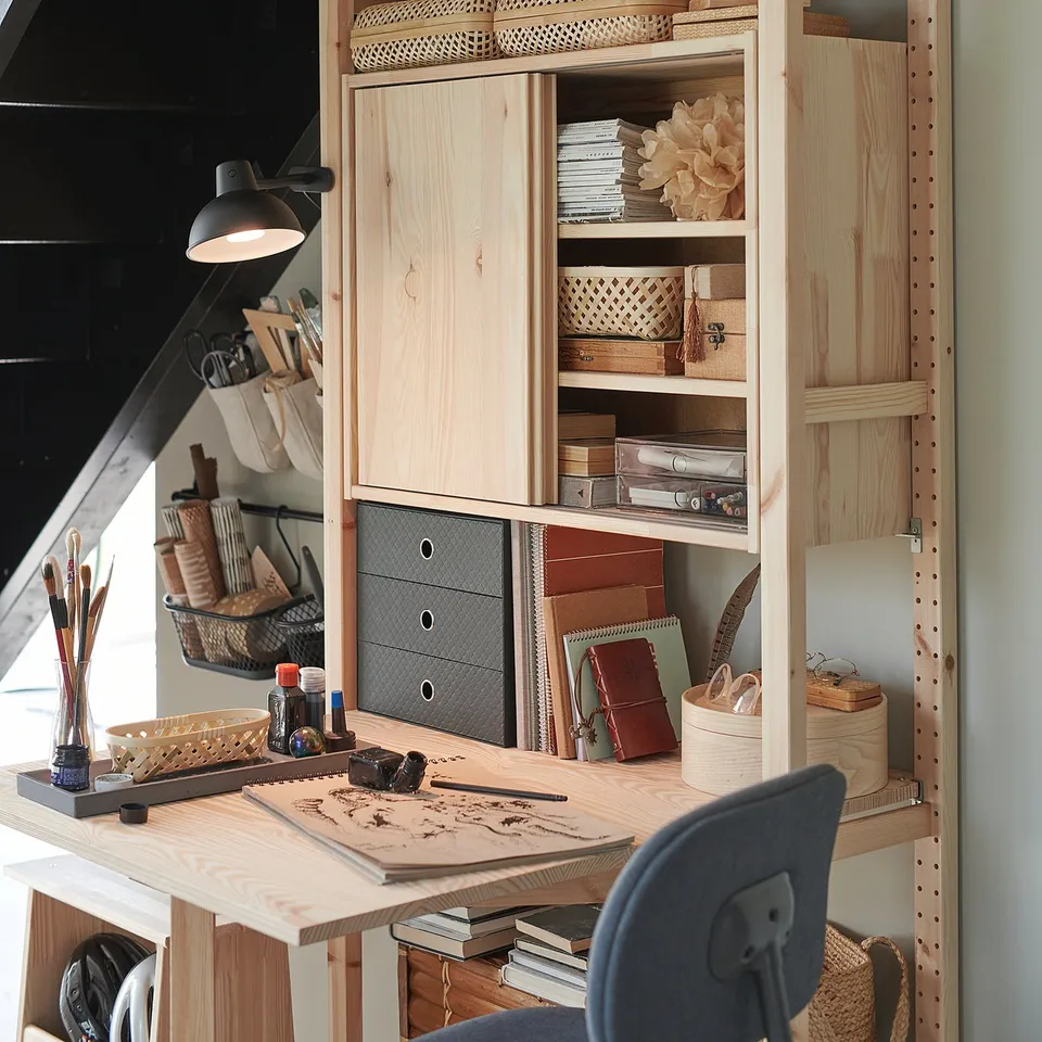 Wooden IVAR desk with shelves, drawers, and various items. Natural finish, containing books, plants, and art supplies.